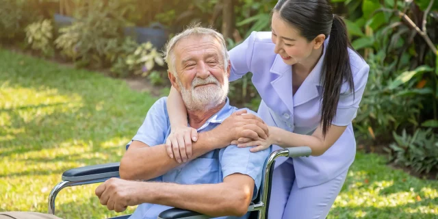 happy-nurse-holding-elderly-man-hand-wheelchair-garden-nursing-home-min